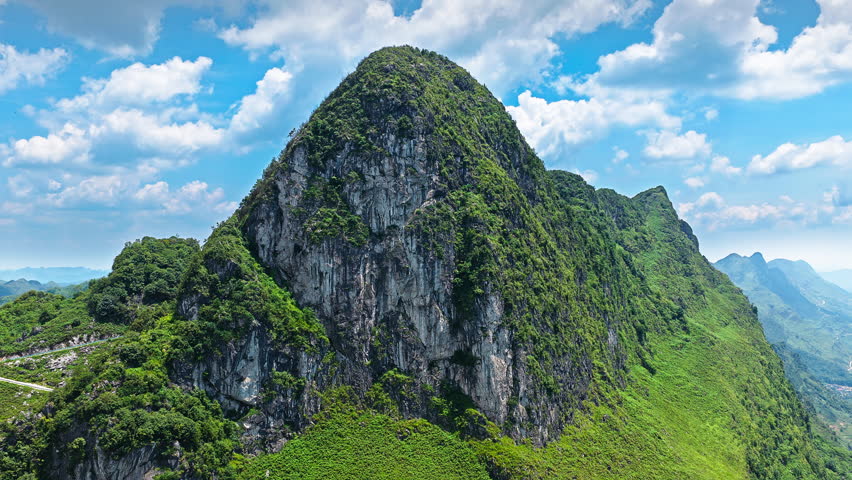 Aerial shot of majestic green karst mountain peak with rocky cliff under blue sky and white clouds