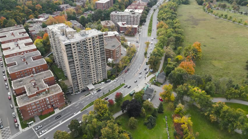 Autumn residential blocks line curving arterial road near Parc La Fontaine in Plateau Mont Royal district as trees show bright seasonal colors captured from clear aerial drone perspective.