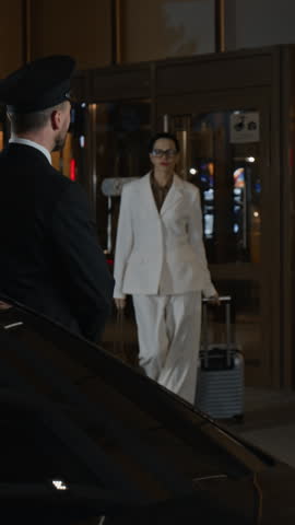 Vertical shot of professional male personal driver in uniform opening car door for woman then helping carrying suitcase while meeting business client at hotel entrance in evening