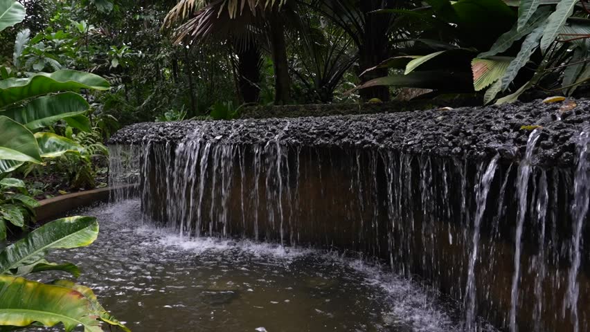 Steadily rising shot of a long fountain system with elevated primary basin providing consistent overflow to a rocky, stone rimmed edge, allowing steady fall of water into large basin below. 