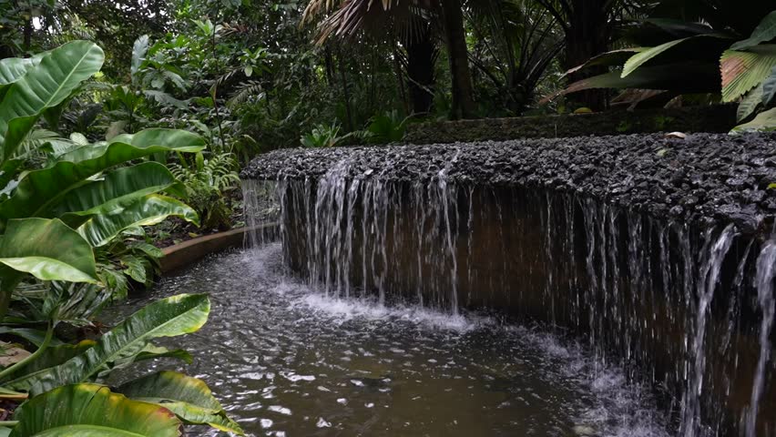 Steady shot of a long fountain system with elevated primary basin providing consistent overflow to a rocky, stone rimmed edge, allowing steady fall of water into large basin below. 
