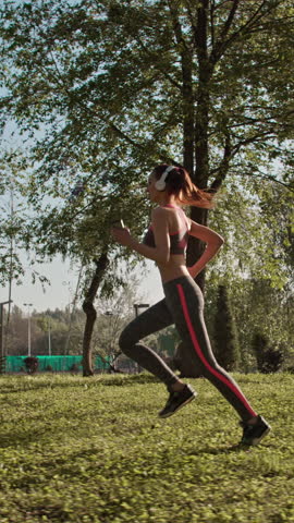 young fit woman running on grass between trees in park listening to music vertical slow motion fullhd healthy lifestyle motivation energy freedom balance outdoor wellness
