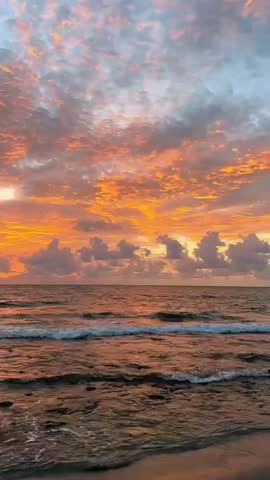 Stunning sunrise over the ocean, with fiery orange and pink clouds glowing above gentle rolling waves that reflect the warm morning light along the peaceful shoreline.