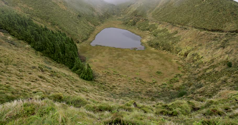 A volcanic lake with clear water in the middle of a crater on a foggy day. The sides of the crater are full of green grass and the sky is cloudy. Tilt up shot.