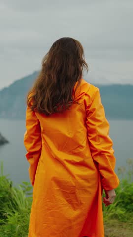 A Person in an Orange Raincoat Stands in Nature, Embracing the Scenic Views Overlooking a Serene Body of Water with Mountains in the Background