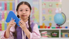 Asian schoolgirl with glasses displays blue number seven, smiling in bright, decorated classroom setting - Powered by Shutterstock - Get 15% off with code: PIKWIZARD15