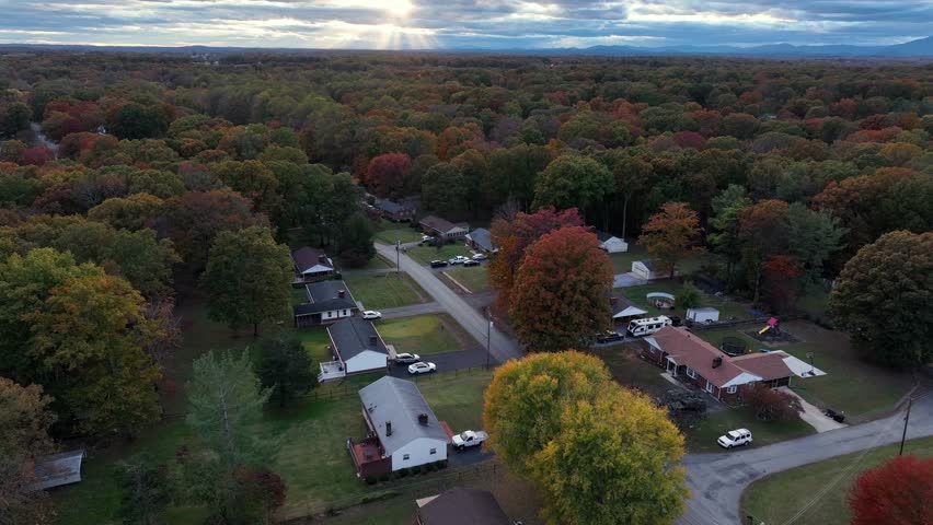 Aerial flyover single level houses in scenic forest landscape of american town. Sunset time behind clouds at horizon. Colored leaves of trees in fall. Wide shot. Serene vibes.