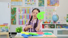 Asian girl holds green foam number six, smiling in brightly lit, colorful classroom, static camera - Powered by Shutterstock - Get 15% off with code: PIKWIZARD15
