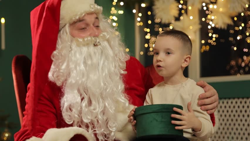 Santa Claus chats kindly with a young boy who holds a green holiday box while sitting near a sparkling Christmas tree