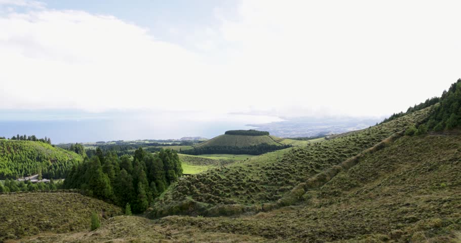 A beautiful landscape with a large hill and the Atlantic Ocean in the background. The hill is covered in trees and grass. The sun is still shining through in Sao Miguel Azores. Trucking shot.