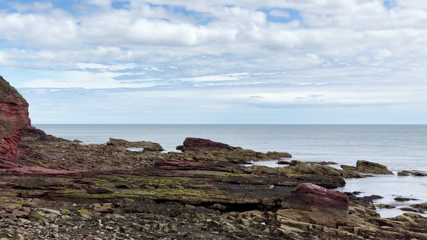 Camera slowly pans over red sandstone cliffs, layered rocks, and North Sea under cloudy daylight