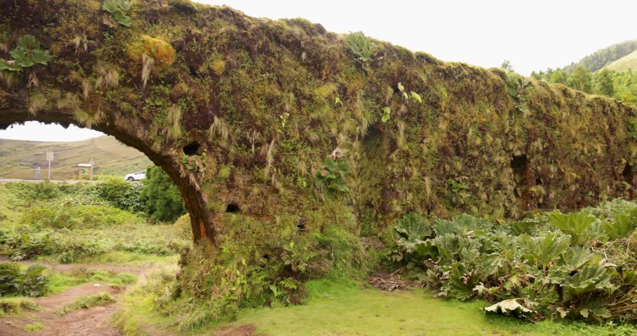 An aqueduct wall and arch with a lush green landscape in the background in Sao Miguel, Azores. The scene is peaceful and serene. Tracking shot.