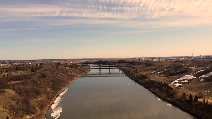 North Saskatchewan River flows toward Edmonton as early sunrise light casts warm tones across calm water and distant bridges creating peaceful prairie edge scene captured from steady drone view.