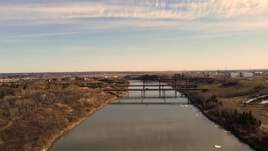 North Saskatchewan River flows toward Edmonton as soft daylight reveals distant bridges, open prairie slopes, and calm water with light ice patches creating wide peaceful scene from steady drone view.