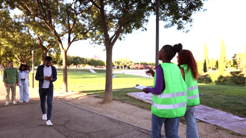 Two dedicated female activists wearing green vests asking people to sign a petition on a clipboard to support their cause, successfully getting signatures from a couple walking in a public park