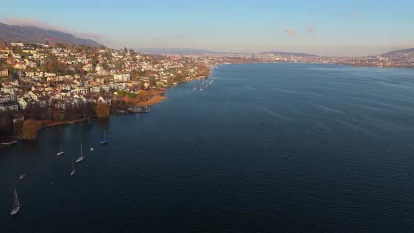 Drone captures wide aerial view from the middle of Lake Zurich. Clear daylight reveals the shoreline of Kilchberg and Zurich city in the distance across calm blue water.
