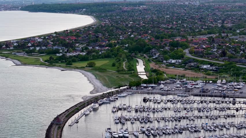 A drone captures a curved breakwater lined with sailboats and a marina on Aarhus Bay, with the city and green coastline visible.