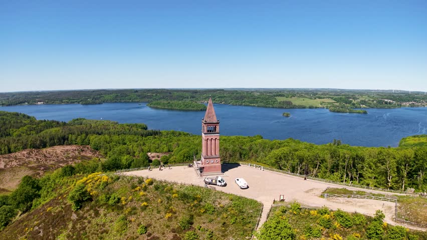 Drone footage captures a historic brick tower standing on a small peninsula by a blue lake in Denmark. The tower and surrounding walkway contrast with lush greenery and calm water.