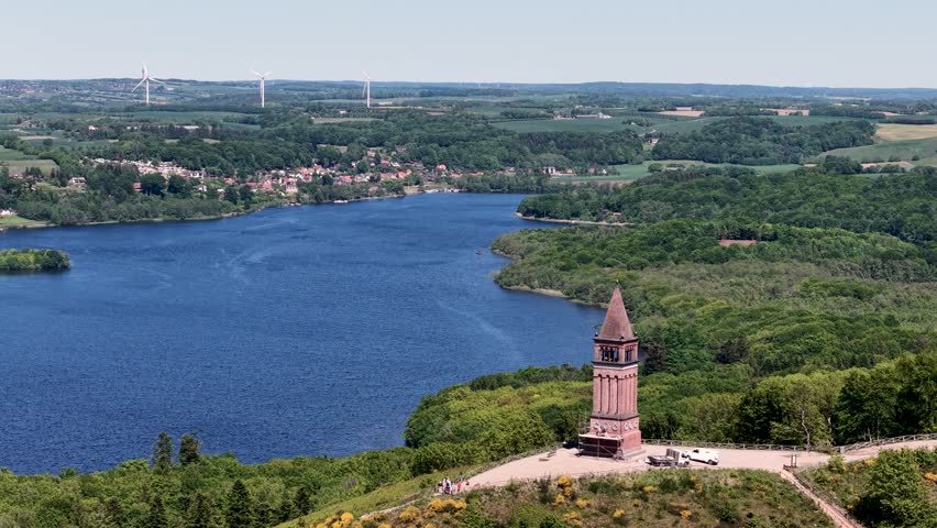 A drone captures the Himmelbjerget tower perched on a hill above a deep blue lake, surrounded by lush forests in Denmark.