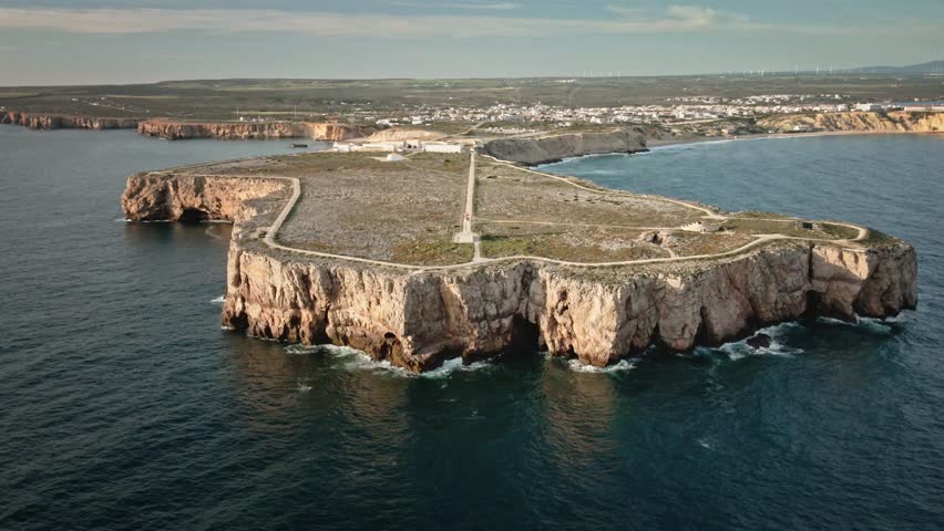 Aerial drone shot moving forward above the ocean, approaching the iconic cliffs, lighthouse, and fortress at the tip of Sagres, Portugal.