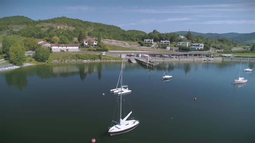 Aerial drone shot tracking sideways across a calm lake with sailboats, a boat launch ramp, and pier at Gasteiz Nautical Club, Ullibarri Gamboa, Spain, on a clear sunny day.