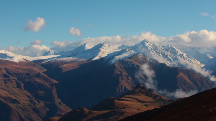 Mountain landscape with snow-capped peaks under blue sky and scattered clouds, rocky slopes in foreground, natural outdoor scenery.
