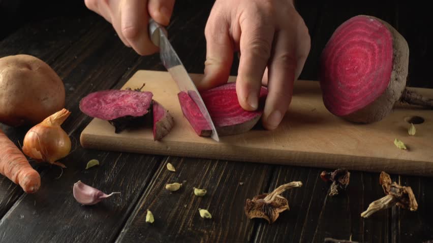 A person uses a sharp knife to cut a fresh purple beet into neat slices on a wooden cutting board. Surrounding ingredients include carrots, garlic, and dried spices.