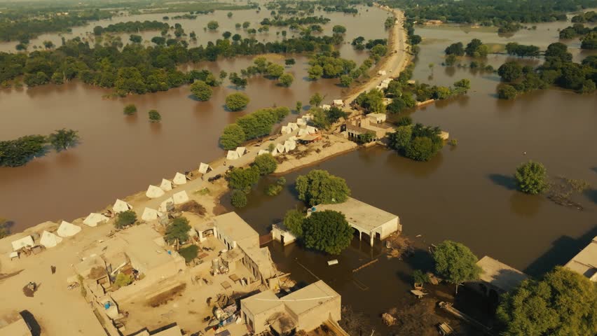 Panoramic pan high above dry residential area showing dirt road lined with tents and flood water on both sides under intense sun.