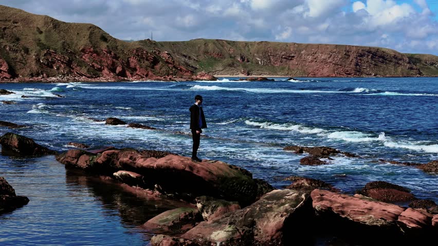 Young man standing on red rock near ocean surf. Teenager watching waves while staying still on coastal stone. Boy observing sea from rocky shoreline. Youth gazing at water while standing quietly on
