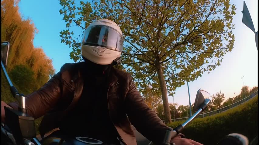 A bottom-angle POV shot of a biker in a white helmet and brown leather jacket riding a silver retro motorcycle beneath a bright blue sky and surrounding trees.
