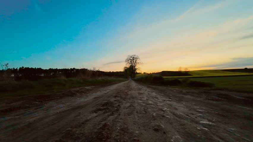 POV car driving on dirt road between fields at sunset. Point of view ride through open countryside with fading golden light. Evening drive along rural path surrounded by farmland and sky