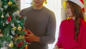 Asian couple wearing santa hat decorates christmas tree with bauble ornament and warm sunlight creating cozy festive mood young woman red sweater and man hanging red ornament christmas tree smiling - Powered by Shutterstock - Get 15% off with code: PIKWIZARD15