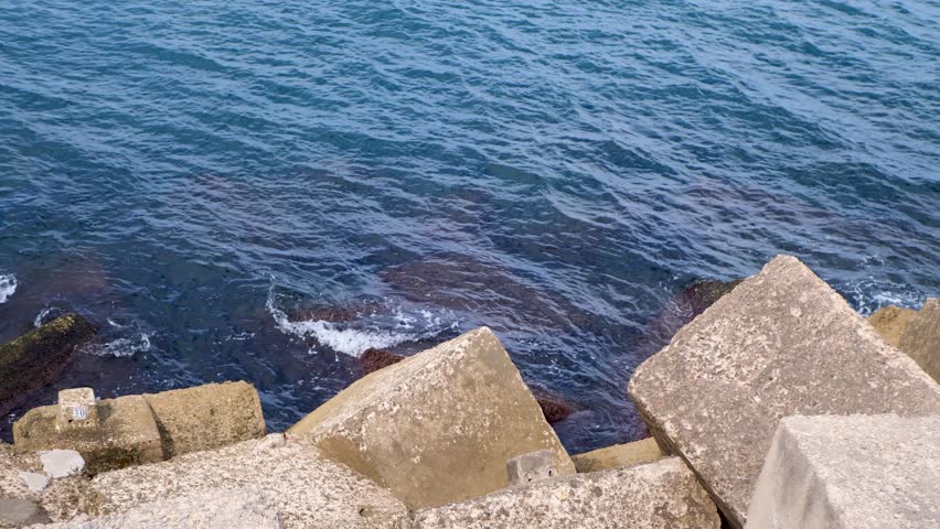 Slow-motion footage of large concrete blocks by shoreline with deep blue sea in background, capturing coastal textures, industrial elements and serene maritime atmosphere - Powered by Shutterstock - Get 15% off with code: PIKWIZARD15