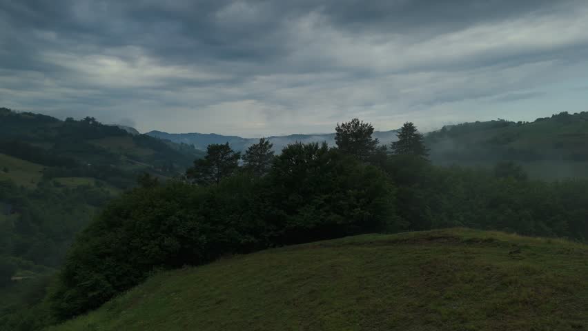 Moody aerial landscape of the Transylvanian hills in Romania, showing a mist filled rural valley, rolling green pastures, hay meadows and scattered farmhouses in the foothills of Carpathian Mountains