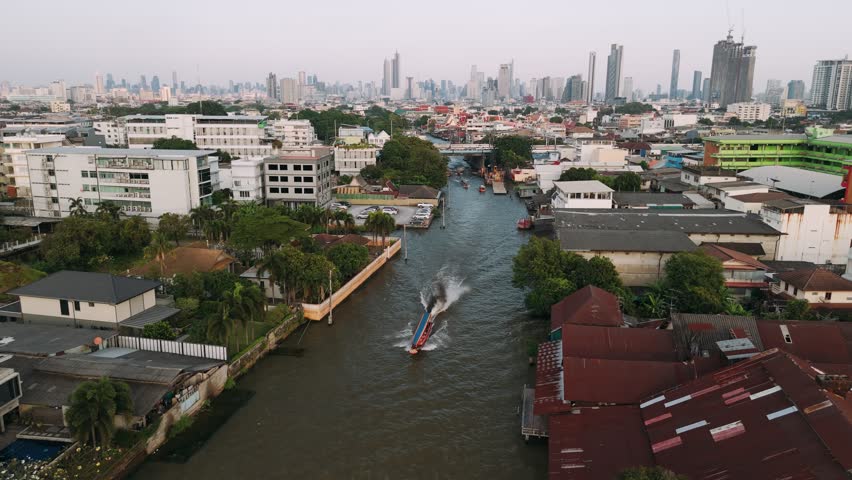 Aerial view of a longtail boat cruising along a bangkok canal, passing by wat paknam phasi charoen temple at sunset, Thailand