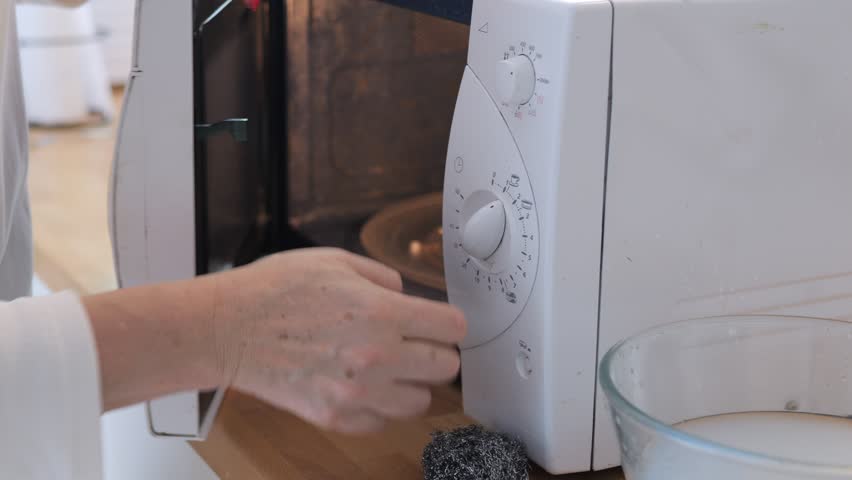 A woman with a red manicure is cleaning the front of the microwave with a sponge.