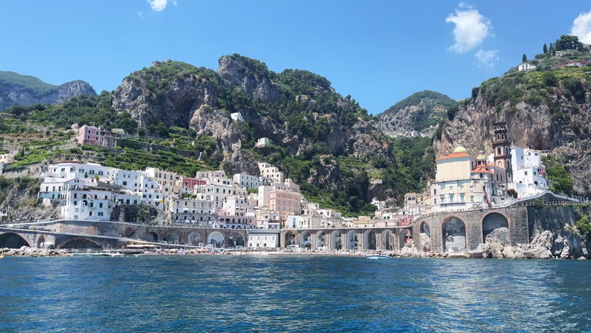 View from sailing boat on beautiful Atrani city located at the foot of powerful hills and greenery, picturesque old buildings, tourists sea beach and blu water waves, charming panoramic view of Atrani