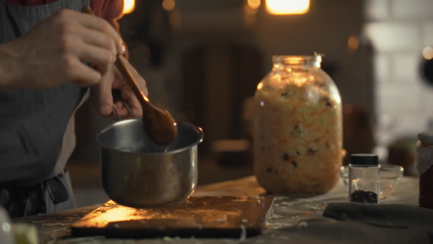 A person stirs rosol in a pot, ready to pour over chopped cabbage for fermentation. This traditional process takes place in a warm kitchen filled with ingredients and tools
