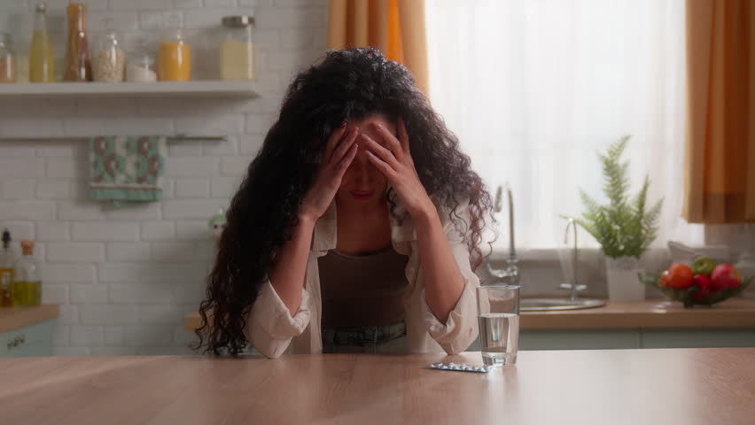 Tired suffering Hispanic Caucasian woman at home kitchen drink pill glass water headache symptom medicine health problem female girl drinking drug indoor ill sick head ache pain painkiller migraine