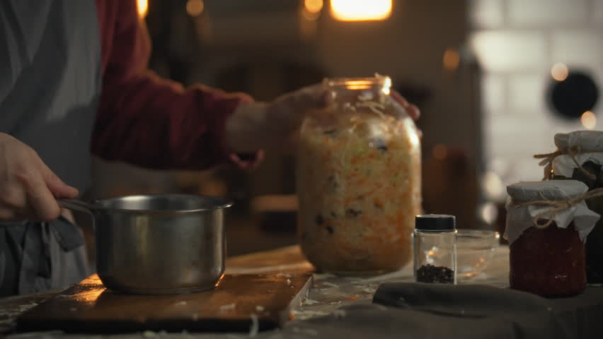 In a warm kitchen, someone pours prepared rossol marinade into a large jar of shredded cabbage to start fermentation. This process is part of making a traditional dish at home