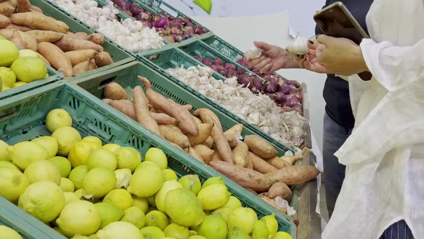 Close-up two women's hands selecting garlic at a market stall.Various vegetables like red onions,long potatoes and lemons displayed in separate boxes.