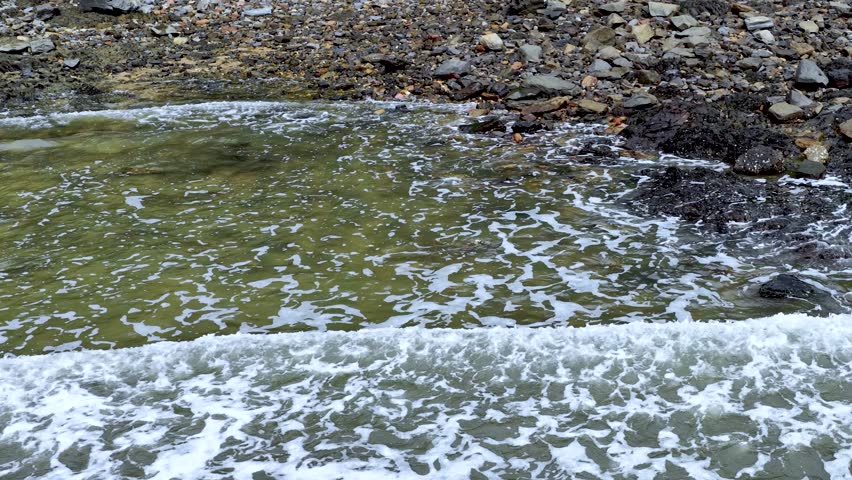 Gentle ocean waves wash over rocky shoreline, foaming water receding, natural daylight, static overhead view