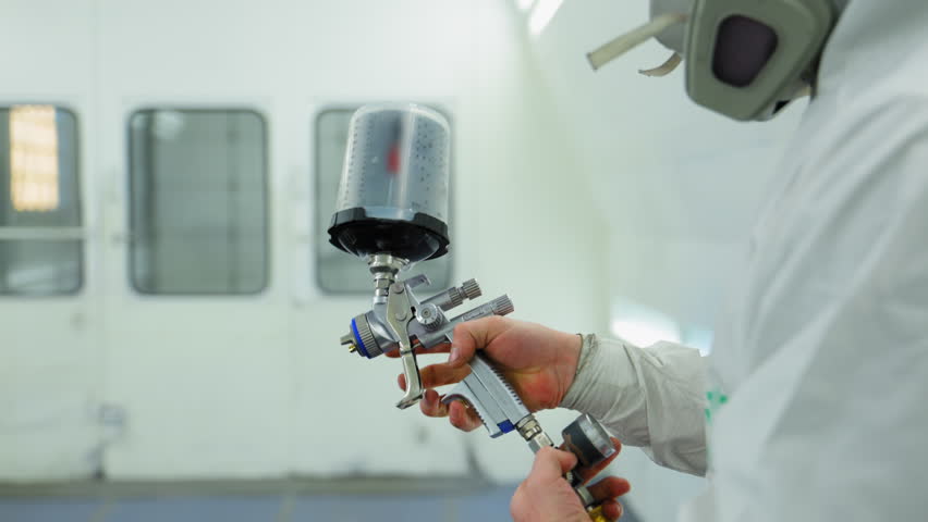 Professional auto body technician test spray gun inside a clean, well lit automotive paint booth. 