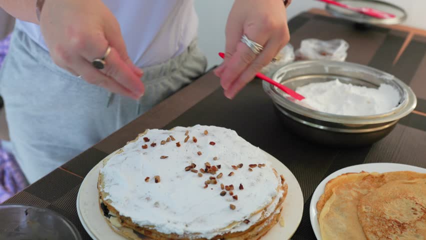 Hands Sprinkling Nuts Over Crepe Cake Layered With Whipped Cream On Table. high angle shot