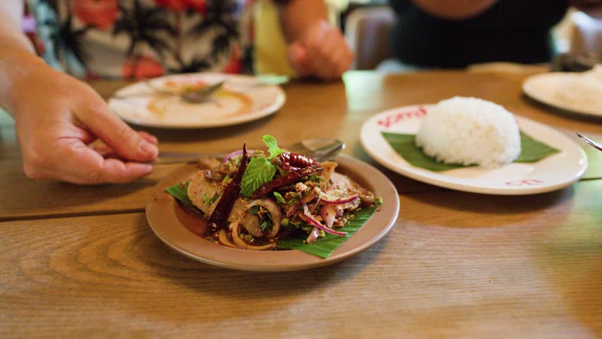 Hand serves spicy pork salad with herbs and rice in warmly lit Bangkok restaurant.
