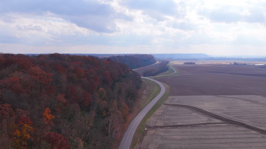 An aerial drone shot of the river bluffs in Southern Illinois. The forest is bright orange with autumn. There is a winding country road running along the edge of the bluffs and surrounding farmland