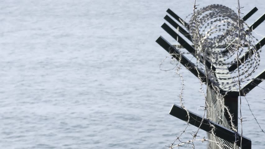 Razor and barbed wire atop a coastal security fence against a gray sea, symbolizing border control, restricted zones, maritime protection, confinement and divided territory