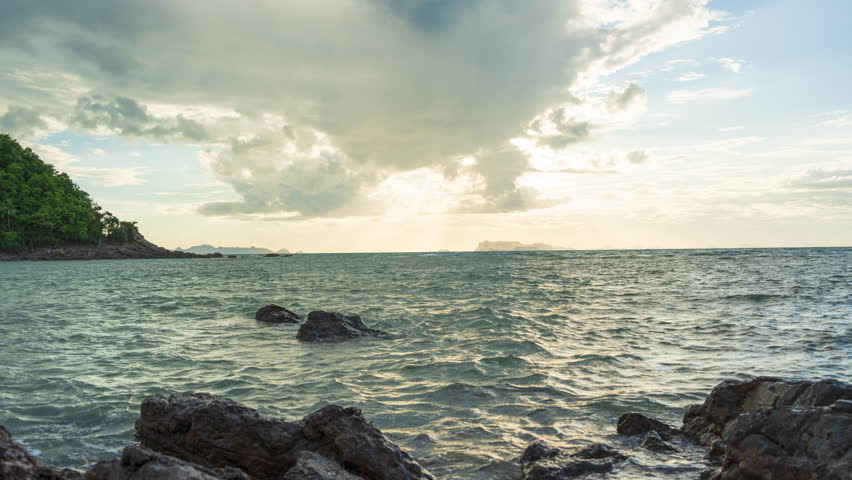 Sun Setting Behind Dramatic Clouds Over Ocean with Rocky Shore in Foreground Captured from Low Angle.