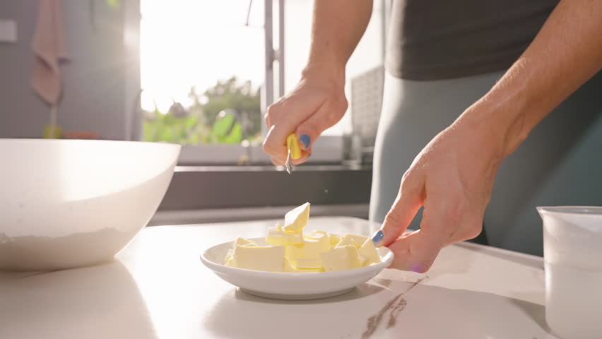 Woman Cutting Butter Cubes on White Plate with Knife near Bowl in Sunlit Kitchen, Close Up Angle, Slow Motion, Baking Ingredient Concept.