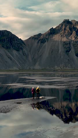 Two hikers stand on a beautiful shoreline at Stokksnes Vestrahorn, surrounded by majestic mountains and serene waters.